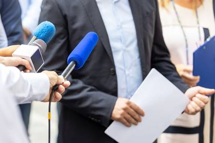 A person in a suit is holding a document and being interviewed by reporters.